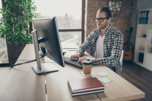 Profissional usando óculos e camisa xadrez sentado ao computador para trabalhar remotamente em seu escritório em casa. 
